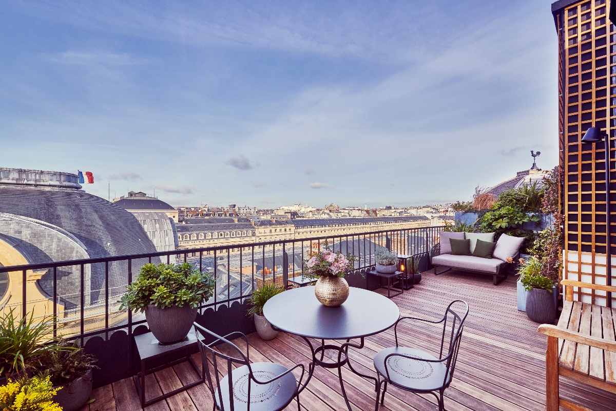 Rooftop terrace with wood decking featuring a dark metal dining table and chairs in the foreground with a vase of flowers, surrounded by potted plants and greenery along the perimeter. The space includes additional outdoor seating with cushioned furniture, a metal safety railing, and a lattice privacy screen on the right side. The terrace overlooks Parisian rooftops and Haussmann-style buildings under a blue sky with light clouds, with a French flag visible on a building to the left.