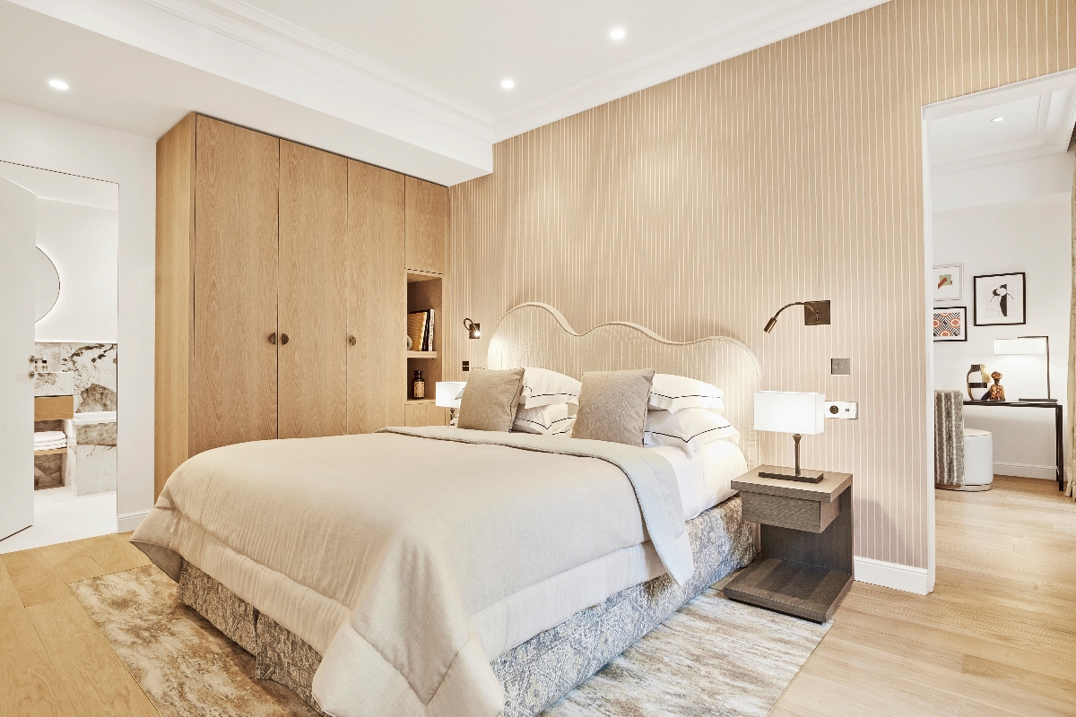 Hotel bedroom featuring a bed with cream-colored linens and a distinctive white upholstered headboard with scalloped edges, positioned against a textured vertical-striped accent wall. The room includes light wood plank flooring, built-in wooden wardrobes on the left wall with integrated shelving, and floating dark wood nightstands with white table lamps and wall-mounted reading lights. Doorways on either side provide views into an adjacent bathroom and connecting room, with framed artwork visible in the space to the right.