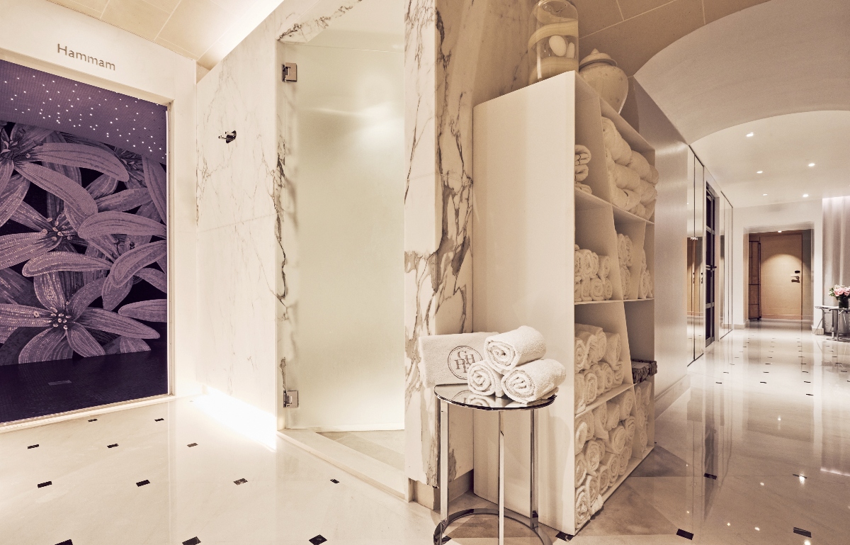 Spa and wellness facility entrance featuring light marble walls with natural veining and white marble flooring with dark diamond inlays. A sculptural display shelf showcases decorative stone vessels and carved architectural elements in cream and beige tones, positioned next to a metal side table holding rolled white towels. The space includes a glass door entrance and a dark purple wall panel with botanical relief artwork labeled "Hammam," with a white-tiled corridor extending into the background.