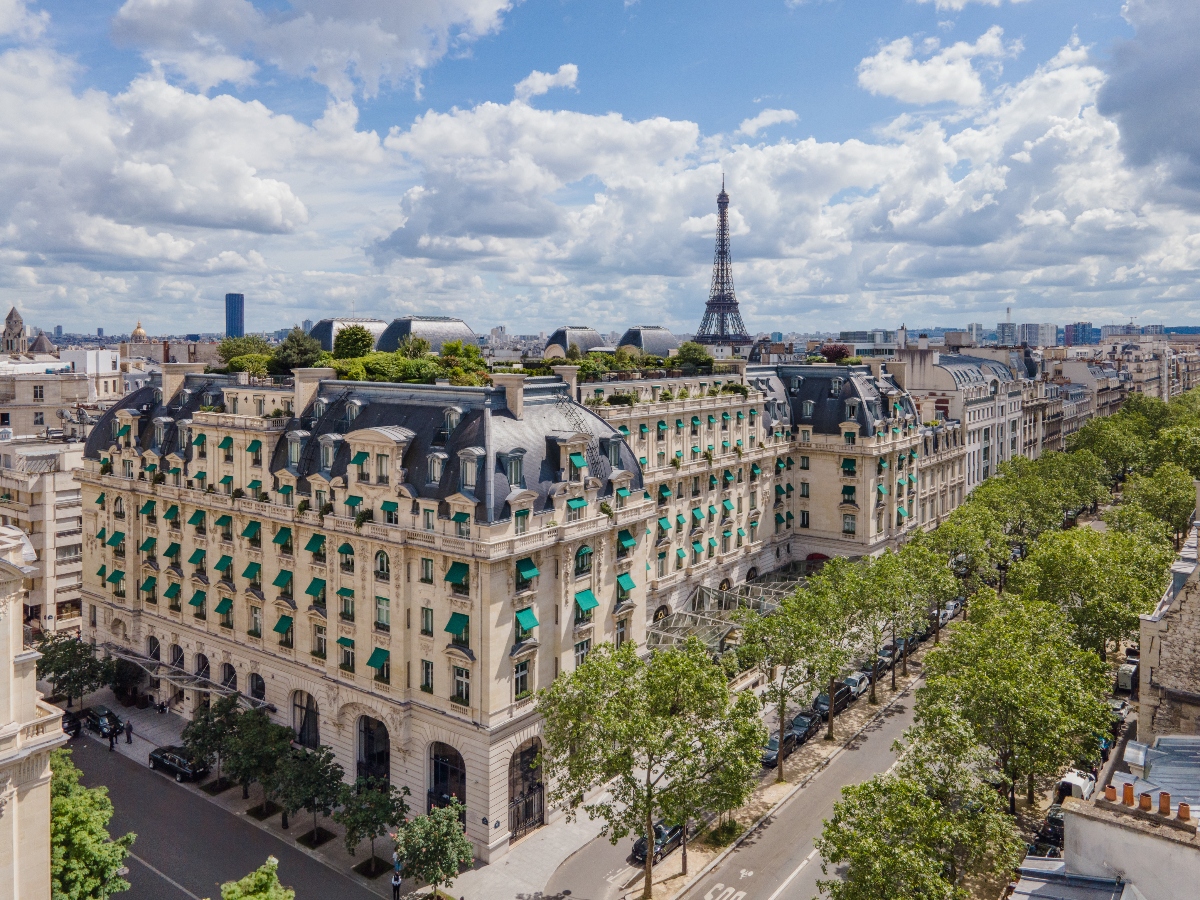 An aerial view captures a classical Parisian building with cream stone facade featuring turquoise awnings at each window across multiple floors, topped with a traditional slate mansard roof. The Eiffel Tower rises prominently in the background against a partly cloudy sky, while tree-lined streets and surrounding Parisian architecture fill the urban landscape. The building sits at a corner location with landscaped surroundings and neighboring historic structures visible throughout the cityscape.