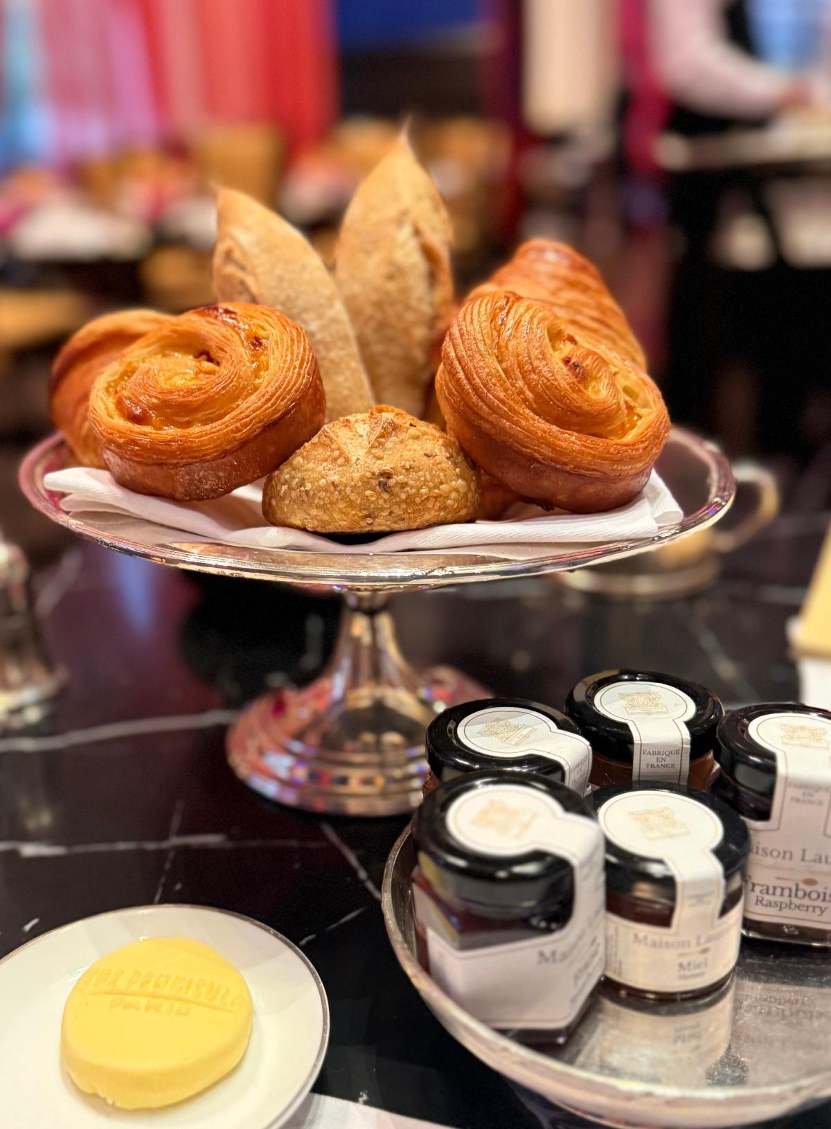 A silver tiered serving stand displays assorted pastries including two golden spiral pastries with visible laminated layers, sliced bread, and a scone arranged on white paper. Below the stand, a small white plate holds a pat of butter, while a silver tray contains multiple small glass jars of preserves with white labels. The breakfast service is set on a dark surface with blurred dining elements visible in the background.
