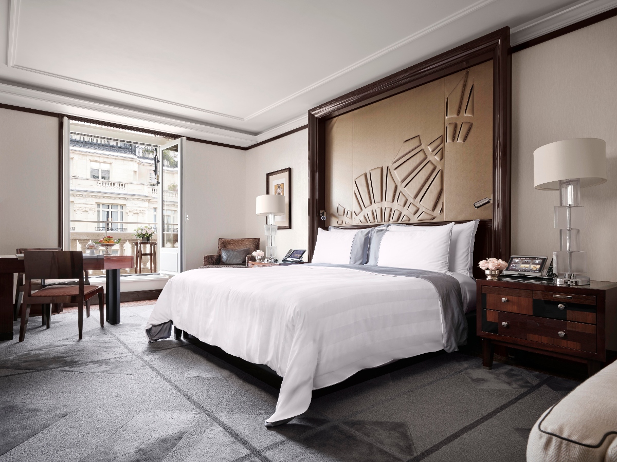 A hotel bedroom features a king bed with white linens and gray accents, centered beneath an Art Deco-inspired headboard with geometric sunburst design in a dark wood frame. French doors open to a balcony overlooking Parisian architecture, while dark wood furnishings include nightstands with crystal lamps, a writing desk with chairs, and a seating area. The room has gray carpeting, white walls with decorative molding, and dark wood trim throughout.