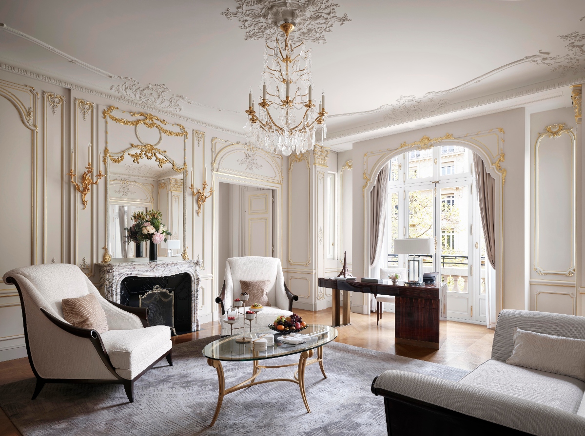 A suite interior features ornate white paneling with gold leaf detailing, elaborate ceiling moldings, and a crystal chandelier suspended from a decorative ceiling medallion. The room includes a white marble fireplace with gold accents, arched French doors opening to a balcony, and upholstered seating arranged around a glass and brass coffee table. Light wood flooring contrasts with the white walls, while a dark wood desk sits near the windowed area draped with floor-length curtains.