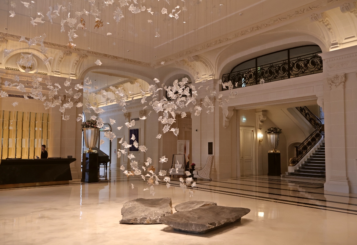 A grand hotel lobby features classical white architecture with ornate moldings, arched doorways, and a sweeping staircase with wrought iron railings. The polished white marble floor is centered by flat stone elements, while an elaborate installation of illuminated white floral branches suspends from the ceiling throughout the space. Reception desk and decorative urns with floral arrangements are visible against the elegant cream and white interior.