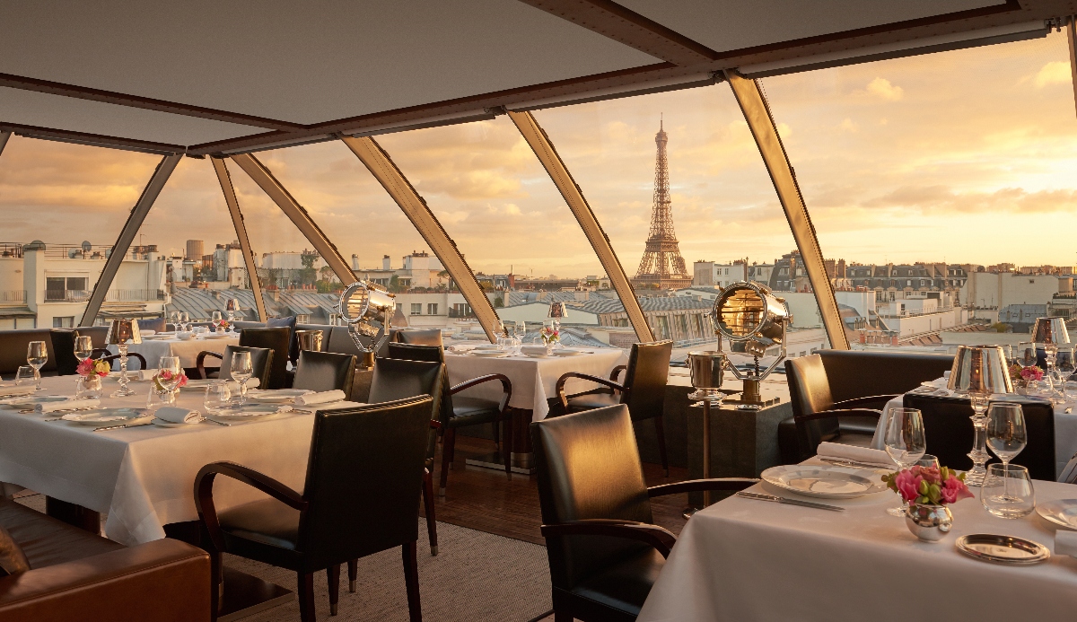 A restaurant dining room features angled floor-to-ceiling windows with geometric metal framing, offering views of Parisian rooftops and the Eiffel Tower under golden evening sky. Tables are set with white linens, wine glasses, place settings, and small floral arrangements, surrounded by dark upholstered dining chairs. The space has gray carpeting and a white ceiling with dark architectural beams that align with the window supports.