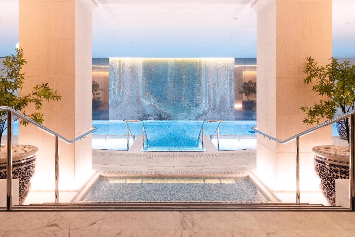 An indoor pool area features cream-colored stone surfaces and square columns, with steps descending into blue water flanked by chrome handrails. A waterfall wall with backlit panels creates cascading water effects in the background, while decorative planters with greenery are positioned symmetrically on either side of the entrance steps. The space has light tile flooring and a minimalist design with warm ambient lighting.