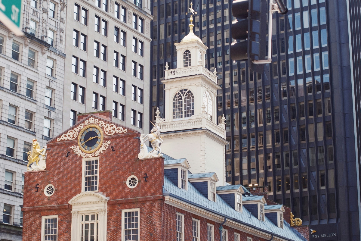 A historic colonial-era brick building with an ornate clock and decorative scrollwork on its facade, topped by a white bell tower with a golden finial, stands surrounded by modern glass and steel skyscrapers. The building features Georgian architectural details including a golden lion statue, white trim, circular windows, and a mansard roof with dormer windows. The stark contrast between the small-scale 18th-century red brick structure and the towering contemporary office buildings behind it illustrates the layered urban development of the cityscape.