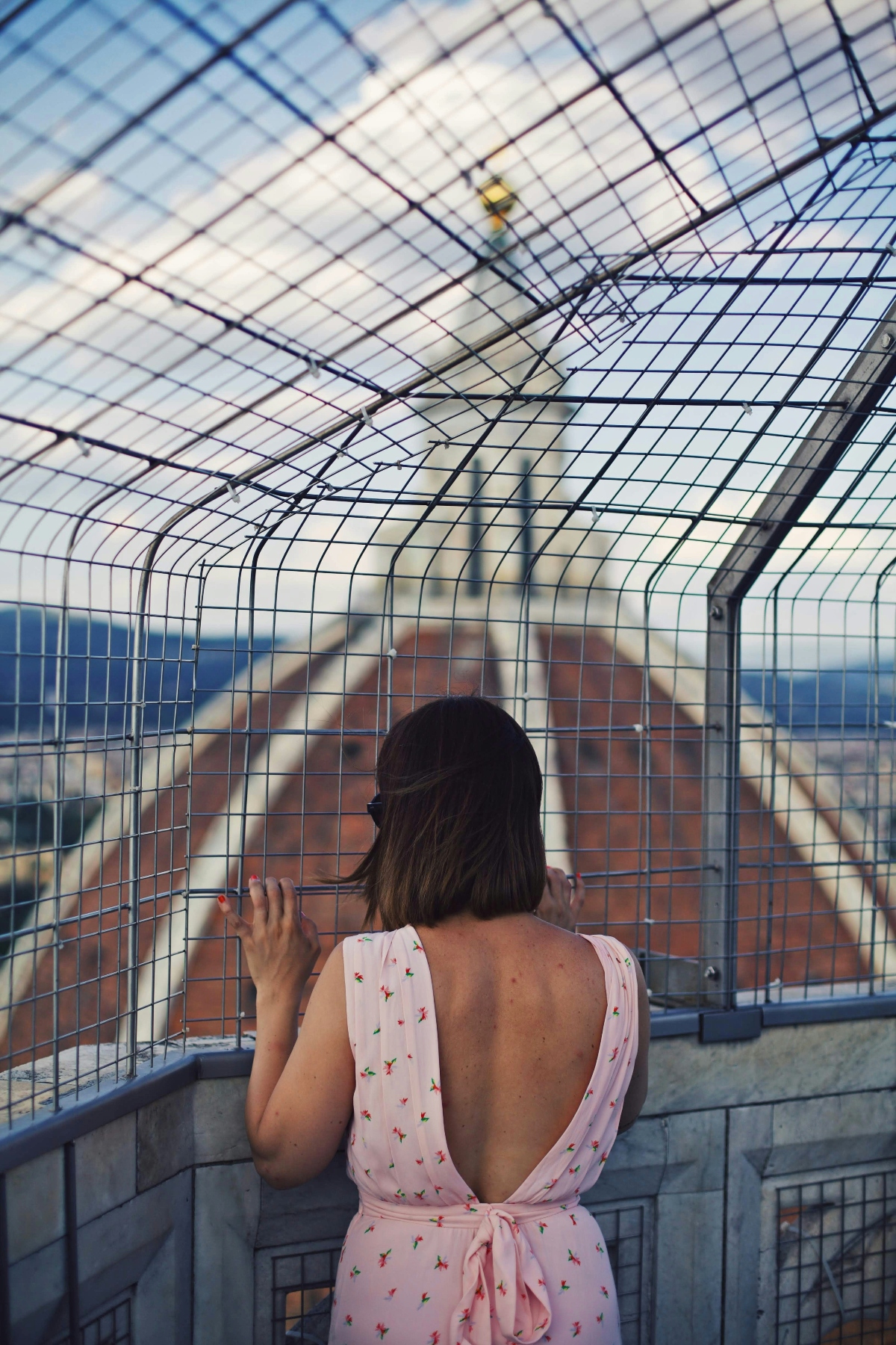 A person wearing a pink floral-print dress with an open back stands at a viewing platform enclosed by curved metal safety barriers, gripping the protective wire mesh while gazing out over a historic cityscape. The view reveals densely packed terracotta-roofed buildings and a prominent Renaissance dome rising in the background against a blue sky. The curved metal grid structure creates a protective cage around the elevated observation point, with the geometric pattern of the barrier framing the panoramic vista.