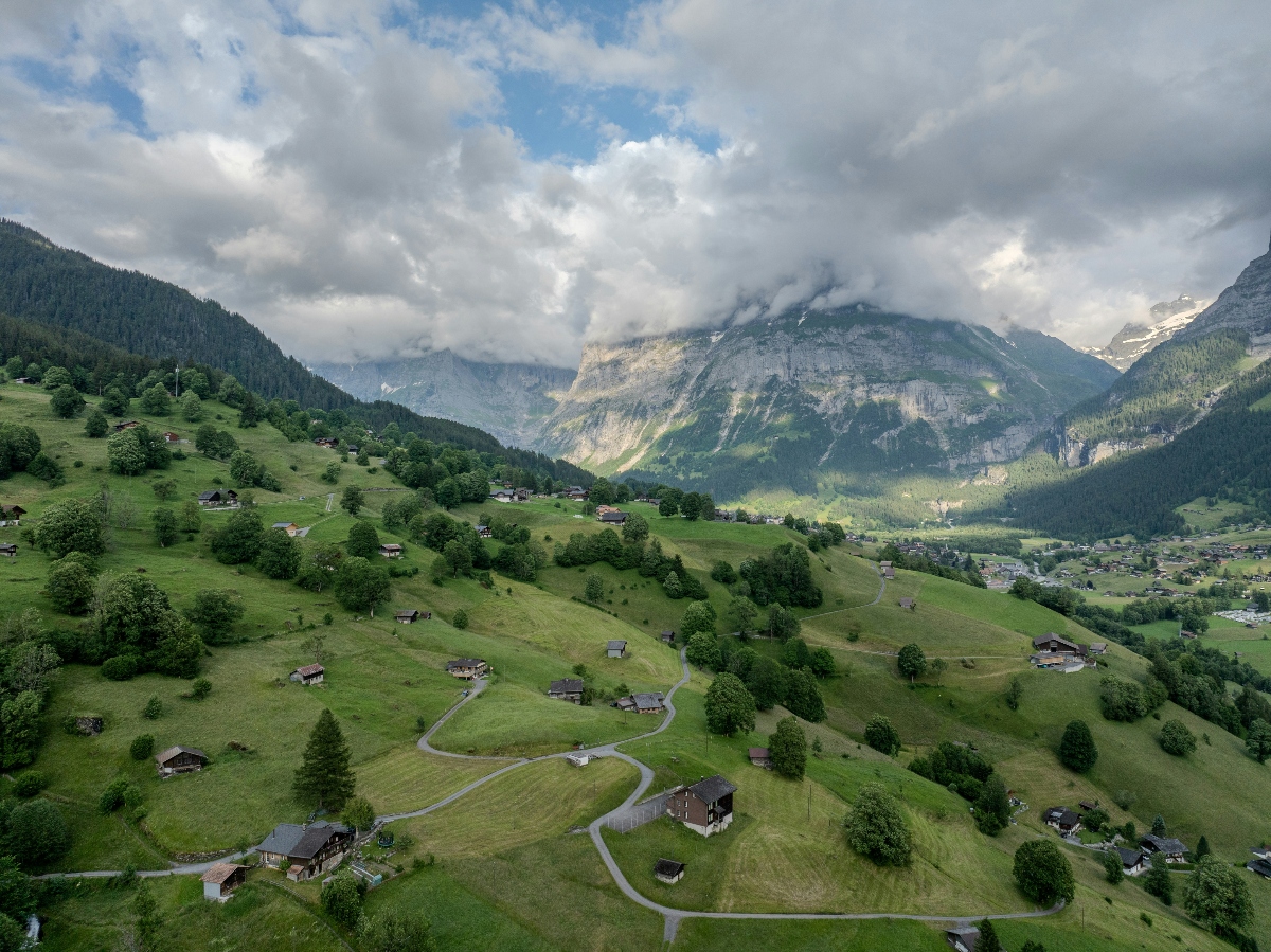 An aerial view of a Swiss Alpine valley with vibrant green meadows dotted with traditional wooden chalets and farm buildings connected by winding roads. In the background, dramatic mountain peaks rise with exposed rock faces, forested slopes, and patches of snow on the higher elevations, partially obscured by dramatic clouds. A village settlement is visible in the valley below, nestled among the rolling hillsides and scattered trees.