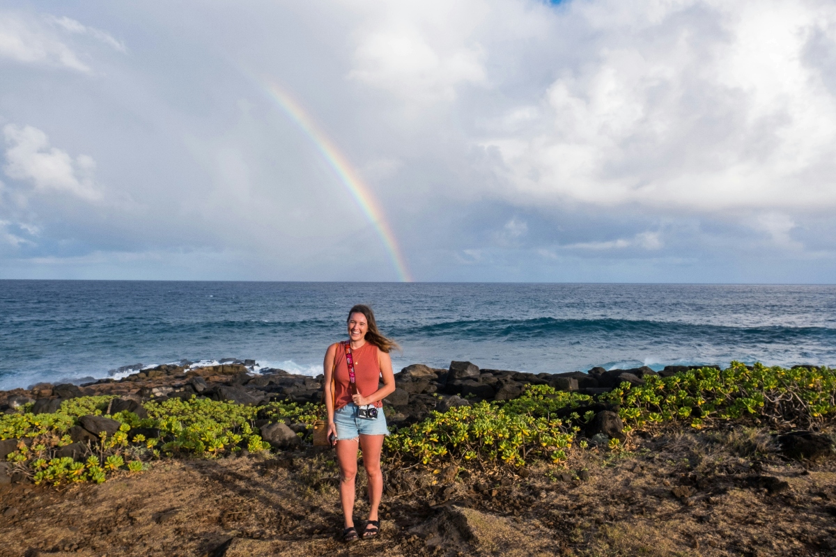 A woman stands on dark volcanic rock terrain holding a camera, wearing a coral-colored tank top and denim shorts, with low-growing green coastal vegetation surrounding the rocky shoreline. Behind her, the deep blue Pacific Ocean meets a dramatic sky featuring a prominent rainbow arcing across gray and white clouds. The landscape is characteristic of Hawaiian coastlines, with black lava rock formations creating a stark contrast against the turquoise ocean waters and breaking waves.