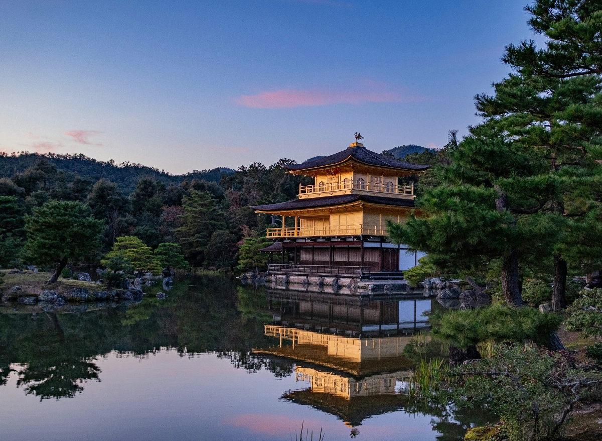 A three-story Japanese pavilion with gleaming golden upper levels and a dark lower level sits at the edge of a tranquil pond, topped by a bronze phoenix ornament and featuring traditional curved roofs with upturned eaves. The structure is perfectly mirrored in the still water, surrounded by meticulously maintained gardens with pine trees, sculpted shrubs, and ornamental rocks, all set against forested hillsides. The scene is captured at twilight with a soft blue and pink sky, while warm lighting illuminates the golden facade of the building.
