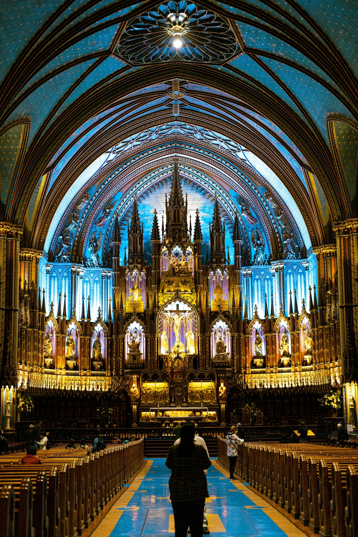 The interior of a Gothic Revival cathedral featuring a soaring vaulted ceiling painted in deep blue with gold stars, supported by dramatic pointed arches with ornate ribbing and decorative patterns. The sanctuary is dominated by an elaborate golden altar screen with multiple Gothic spires, niches containing statues, and intricate carved details, dramatically illuminated by blue and gold lighting. A silhouetted figure stands in the central aisle between rows of wooden pews, facing the altar, providing scale to the monumental architecture.