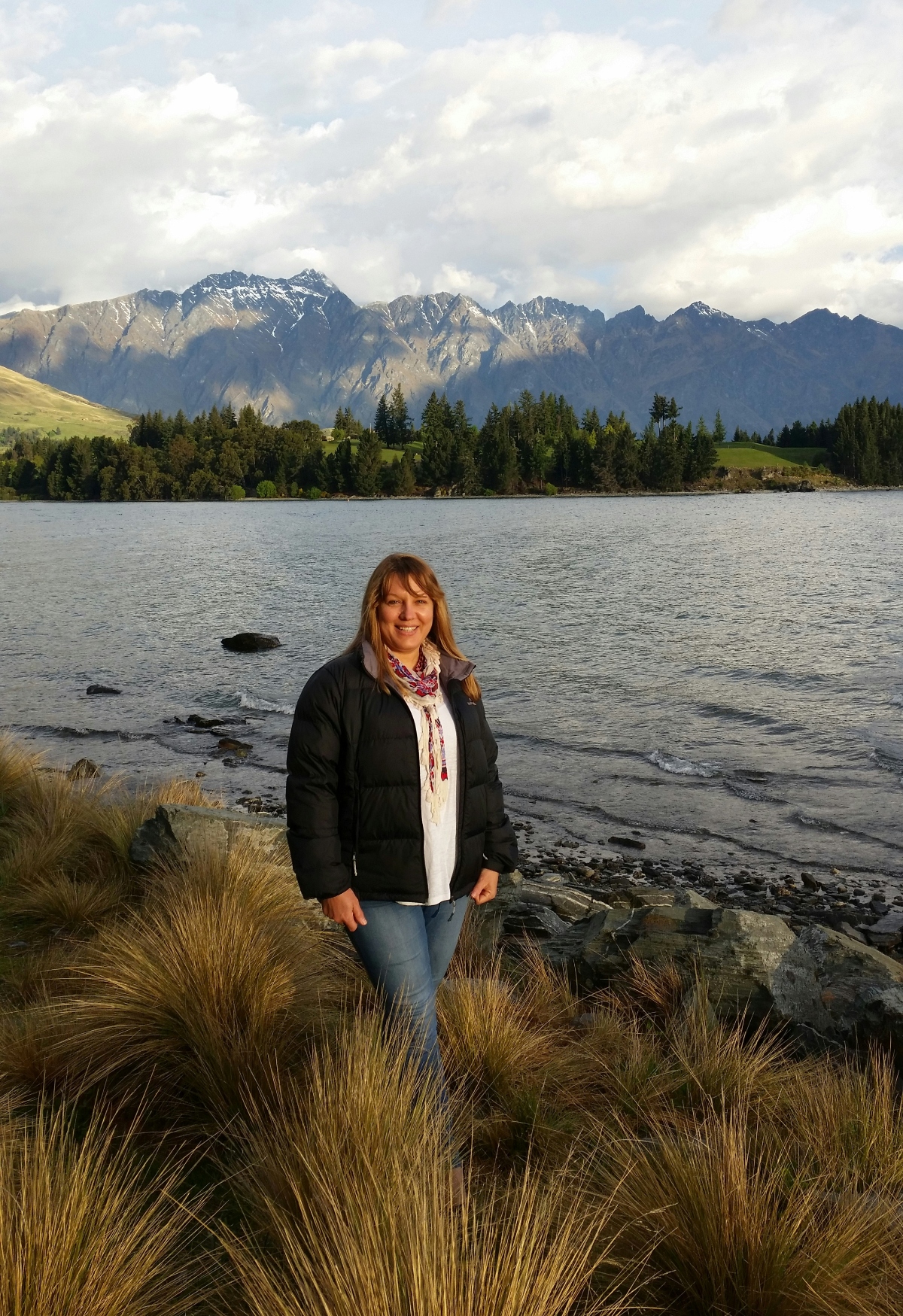 A person wearing a black jacket and patterned scarf stands among golden tussock grasses on a rocky lakeshore, with calm gray-blue water stretching behind them toward a tree-lined opposite shore. Beyond the dense evergreen forest, a dramatic mountain range with jagged peaks and patches of snow rises against a cloudy sky. Dark rocks are scattered along the water's edge, and gentle ripples disturb the lake's surface in the foreground.