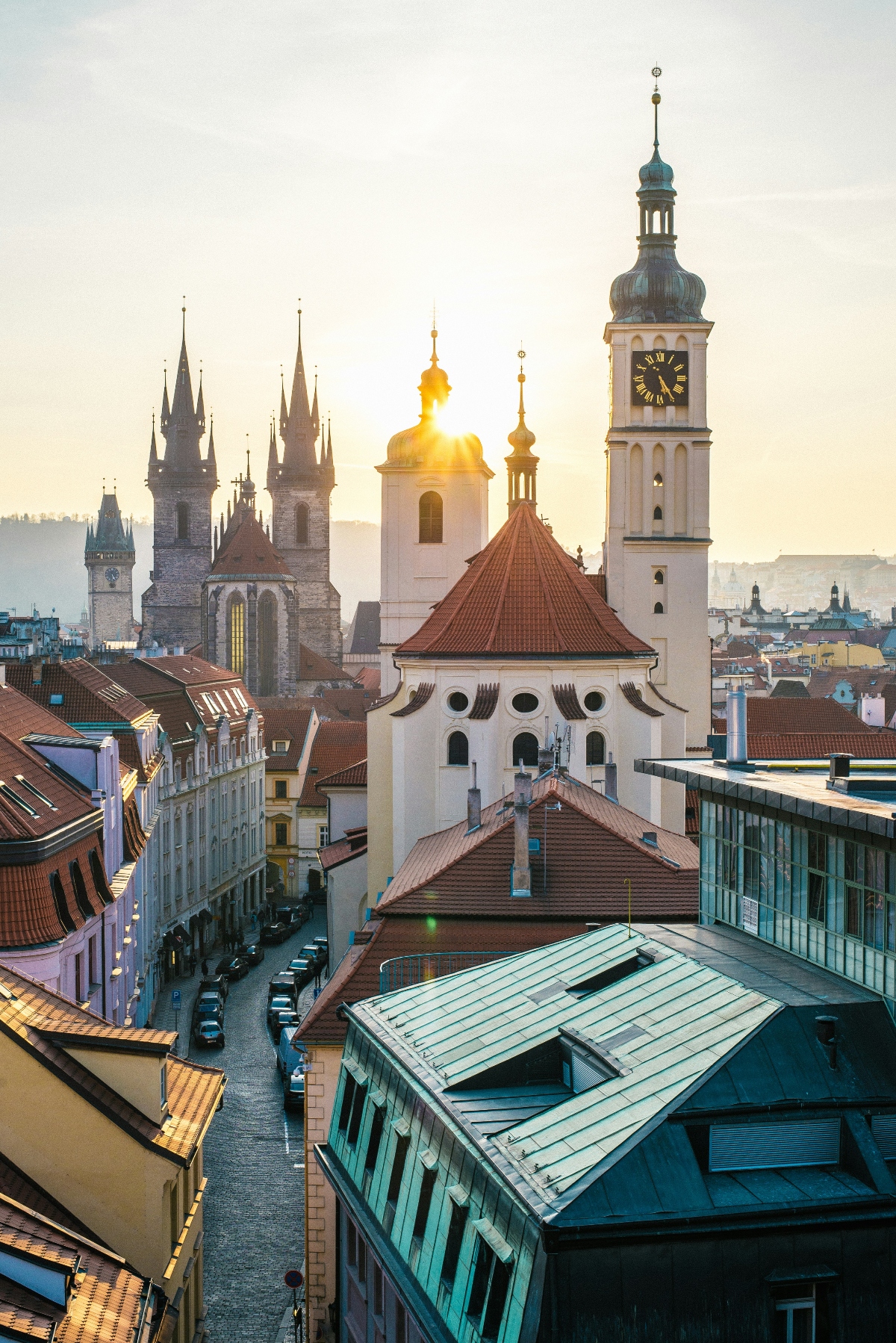 An elevated view of Prague's historic Old Town skyline at sunset, featuring multiple church spires and towers including prominent Gothic twin spires in the background, a white church with a distinctive red conical roof in the foreground, and a tall clock tower with a green copper dome on the right. The cityscape is characterized by densely packed buildings with terra cotta and red tile roofs, narrow streets winding between structures, and warm golden light illuminating the architectural details. The composition captures the layered medieval and baroque architecture that defines Prague's historic center.