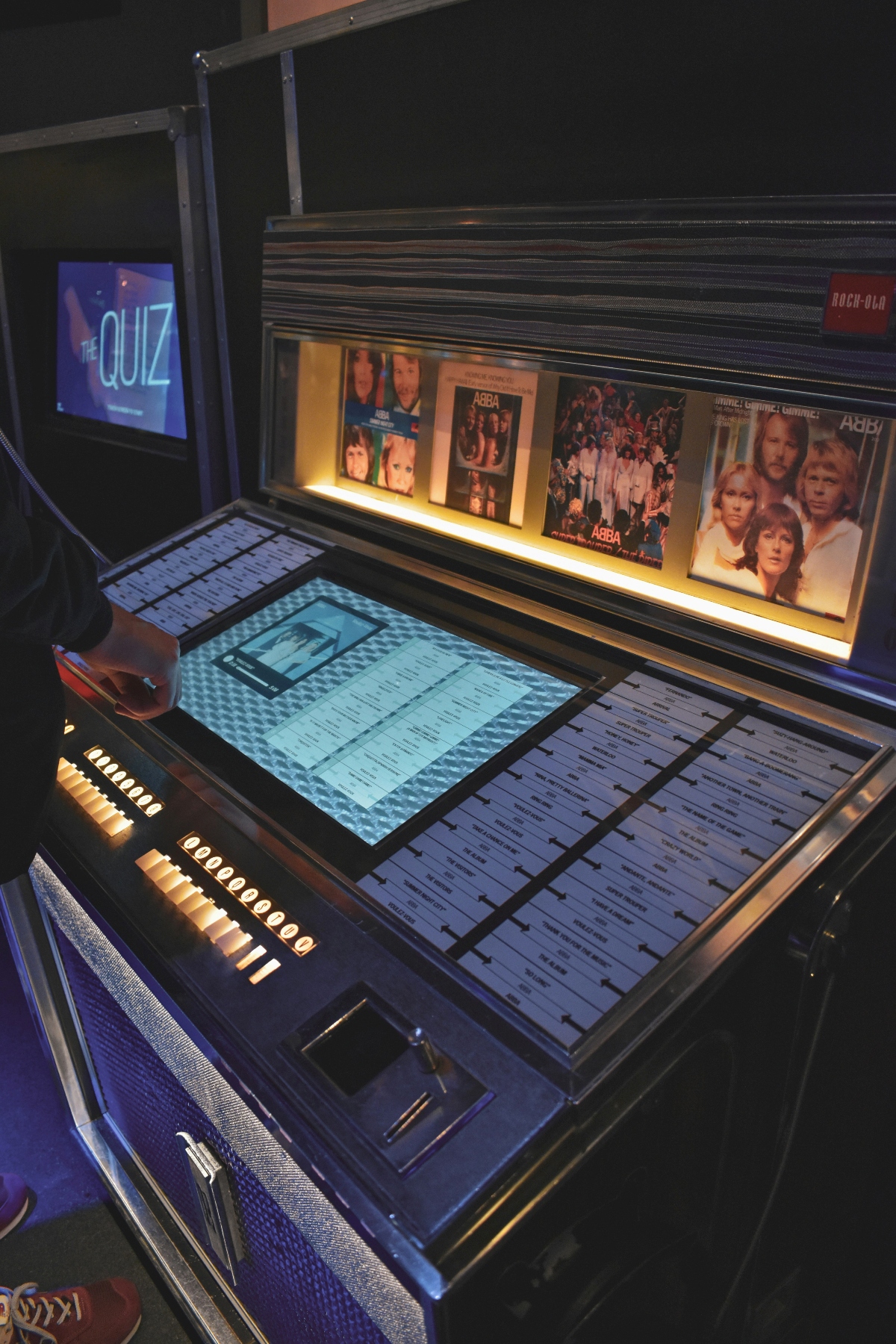 A vintage-style jukebox display with illuminated ABBA album covers visible in the upper section and an interactive touchscreen panel below showing song selections. A monitor displaying "The Quiz" in neon blue lettering is positioned to the left of the jukebox in the darkened exhibition space.