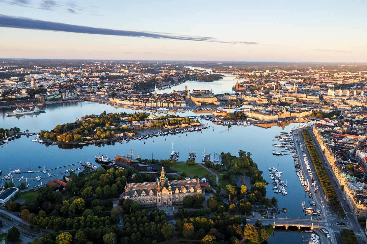 Stockholm's Gamla Stan (Old Town) viewed from across the water at dusk, with historic ochre and cream-colored buildings lining the waterfront and the distinctive spire of Riddarholmen Church rising prominently in the center of the skyline. The sky displays soft purple, pink, and orange tones with scattered clouds, while the calm water reflects the city lights in the foreground.