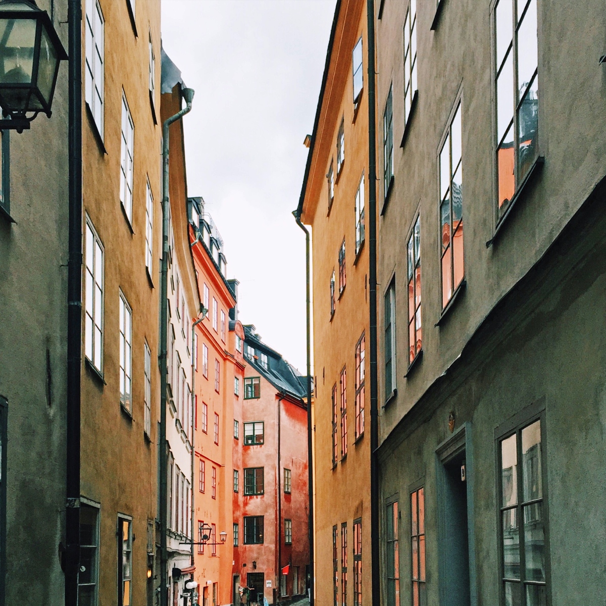 A narrow cobblestone alley in Stockholm's Old Town lined with tall historic buildings in shades of ochre, coral, gray, and orange, their facades showing weathered plaster and regularly spaced windows. The tight medieval street creates a vertical corridor effect, with dark metal lanterns mounted on the walls and a single pedestrian visible in the distance at street level.