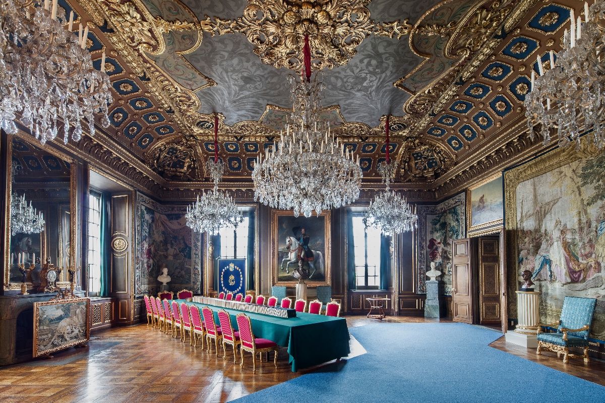 An ornate palace state room featuring an elaborately decorated ceiling with gold leaf ornamentation, gray painted panels with blue medallions, and three massive crystal chandeliers. A long turquoise-draped table is surrounded by rows of pink-cushioned gilt chairs, while the walls display large historical paintings in gilded frames, decorative tapestries, and period furniture on polished parquet flooring covered partially by a blue carpet runner.