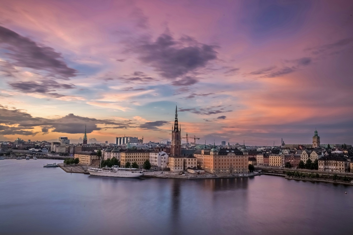 Stockholm's Gamla Stan (Old Town) viewed from across the water at dusk, with historic ochre and cream-colored buildings lining the waterfront and the distinctive spire of Riddarholmen Church rising prominently in the center of the skyline. The sky displays soft purple, pink, and orange tones with scattered clouds, while the calm water reflects the city lights in the foreground.