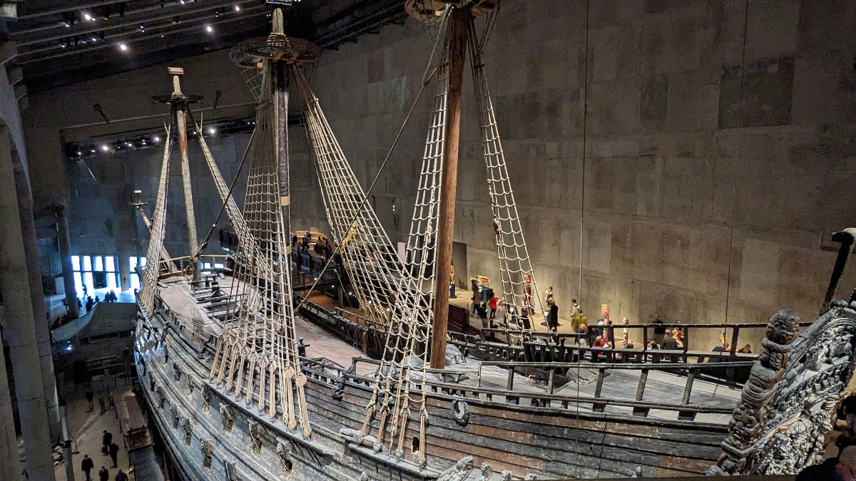The preserved 17th-century warship Vasa displayed inside a purpose-built museum, showing the vessel's dark weathered wooden hull, three tall masts with rigging, and ornately carved stern section. Museum visitors stand on elevated viewing platforms at multiple levels surrounding the ship, which is housed within a large hall with gray concrete walls and overhead lighting.