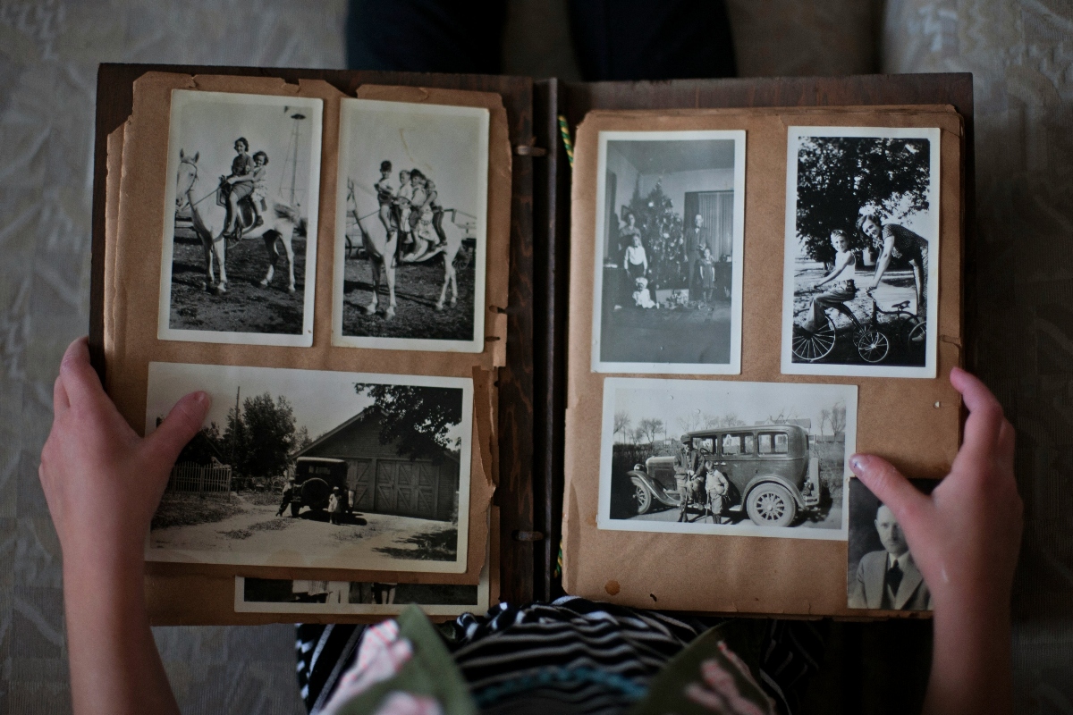 Hands hold open a vintage photo album with brown pages displaying six mounted black-and-white photographs depicting scenes from what appears to be the early-to-mid 20th century, including images of people on horses, a group portrait, a cyclist, a house with a vintage car, and an old automobile with passengers. The worn album pages show signs of age, and the photographs are arranged in a traditional scrapbook layout with visible mounting corners.