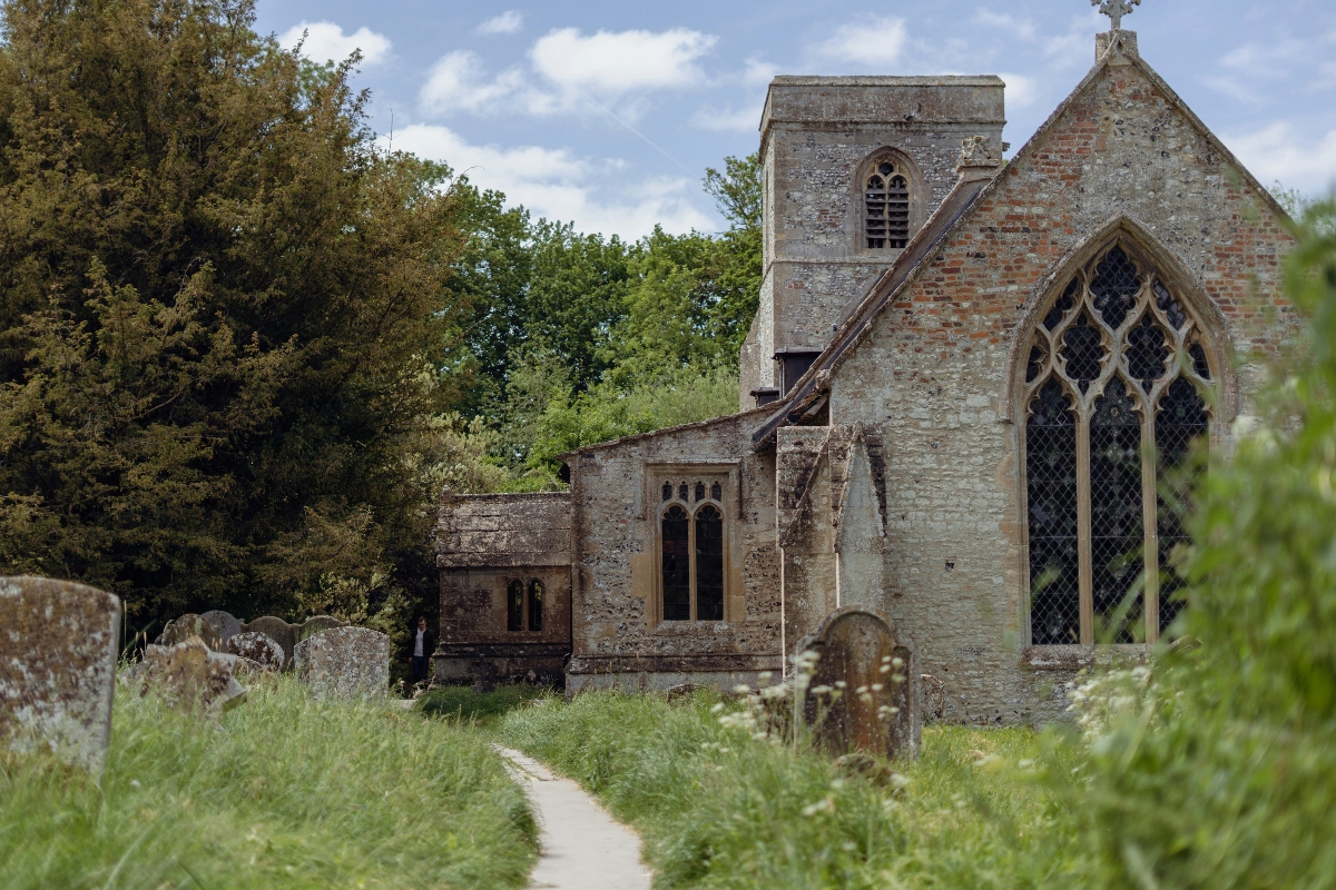 A weathered stone church with Gothic pointed-arch windows and a square tower stands among old gravestones in an overgrown churchyard, with a narrow path cutting through the tall grass. The building displays aged stonework with exposed brick sections, surrounded by dense trees under a partly cloudy sky.