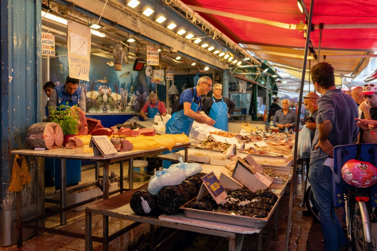 Vendors in blue aprons work behind a seafood stall at an outdoor market, with fresh fish displayed on metal tables and shellfish arranged in containers beneath a red-striped awning. Customers gather around the weathered blue-painted stall structure, which features hanging lights and handwritten price signs, while bicycles and other market activity are visible in the background.