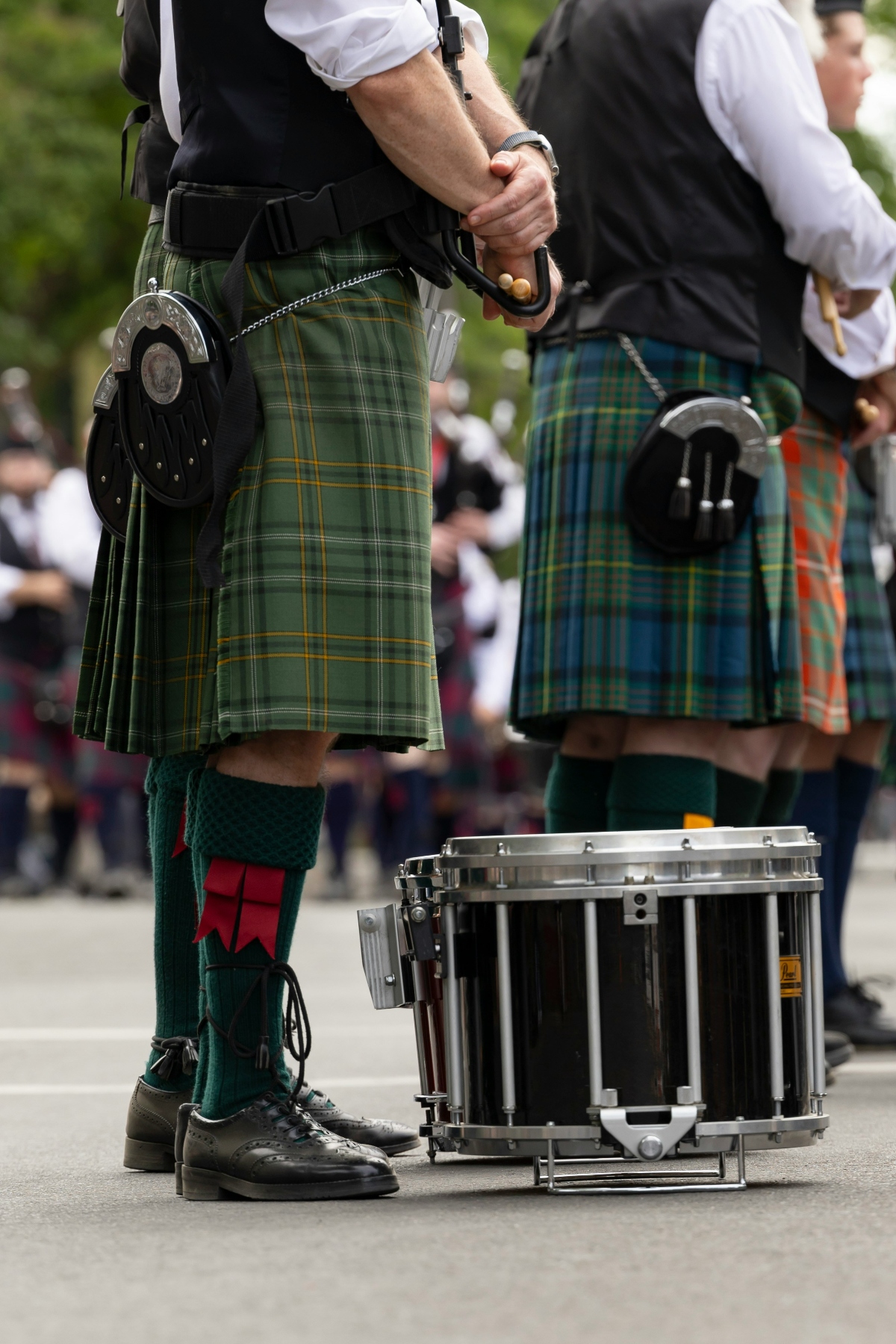 Two bagpipe band members wearing traditional Scottish dress stand in formation, with green tartan kilts, black vests, white shirts, green knee-high socks with red flashes, and sporrans with silver detailing visible at their waists. A silver-rimmed marching snare drum rests on the pavement beside black formal shoes, while the musicians hold drumsticks, with additional band members in similar attire visible in the blurred background.