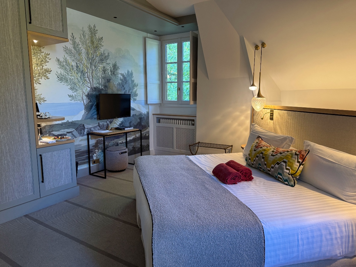 Modern hotel bedroom with white bed featuring woven rattan headboard, gray runner, burgundy towels, and patterned accent pillow, flanked by wall-mounted sconces beneath a sloped ceiling. The room includes a large photographic wall mural depicting a Mediterranean coastal landscape, white window shutters opening to greenery, built-in cabinetry, and light wood flooring.