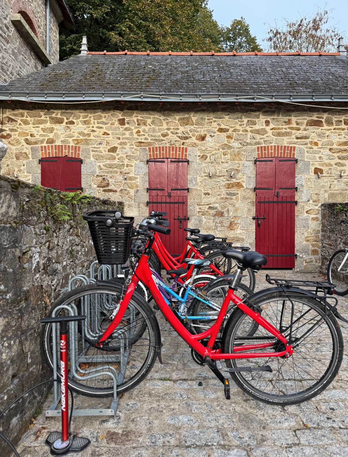 Red and turquoise bicycles parked in metal bike racks on a cobblestone courtyard, with a red-colored bike pump station standing to the left. Behind the bikes stands a historic stone building with three burgundy wooden doors topped by red brick archways, a moss-covered slate roof, and trees visible above.