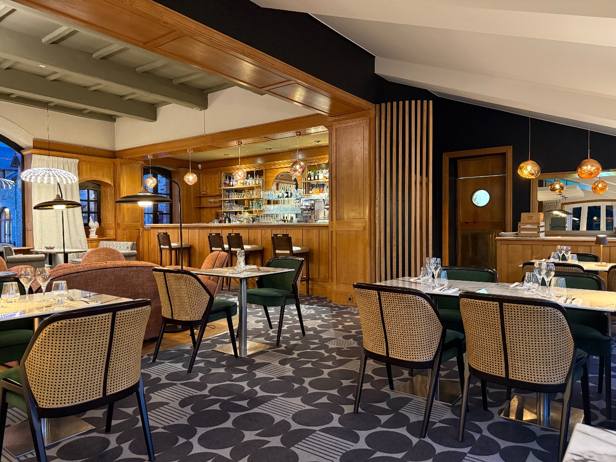 Restaurant dining room featuring tables with dark green velvet chairs and natural cane-back chairs arranged on geometric black and gray patterned flooring, with amber globe pendant lights suspended from the ceiling. The space includes a wooden bar with stools in the background, wood paneling, a black accent wall with vertical wood slat divider, exposed white ceiling beams, and arched windows.