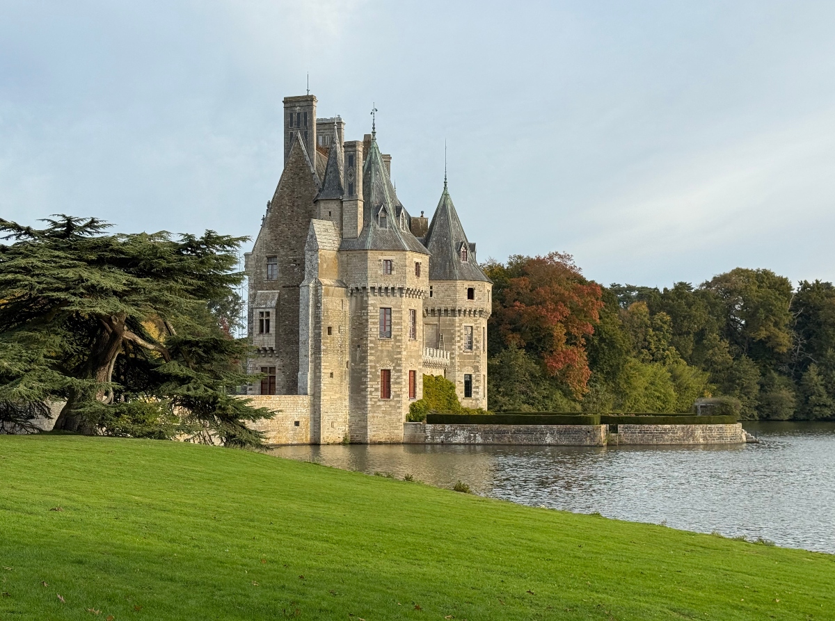 Medieval-style stone château with multiple towers topped by pointed slate roofs and a copper-green spire, situated at the edge of a lake or moat with a stone retaining wall. The building is surrounded by manicured green lawn in the foreground and mature trees including a large conifer and autumn-colored deciduous trees, under an overcast sky.