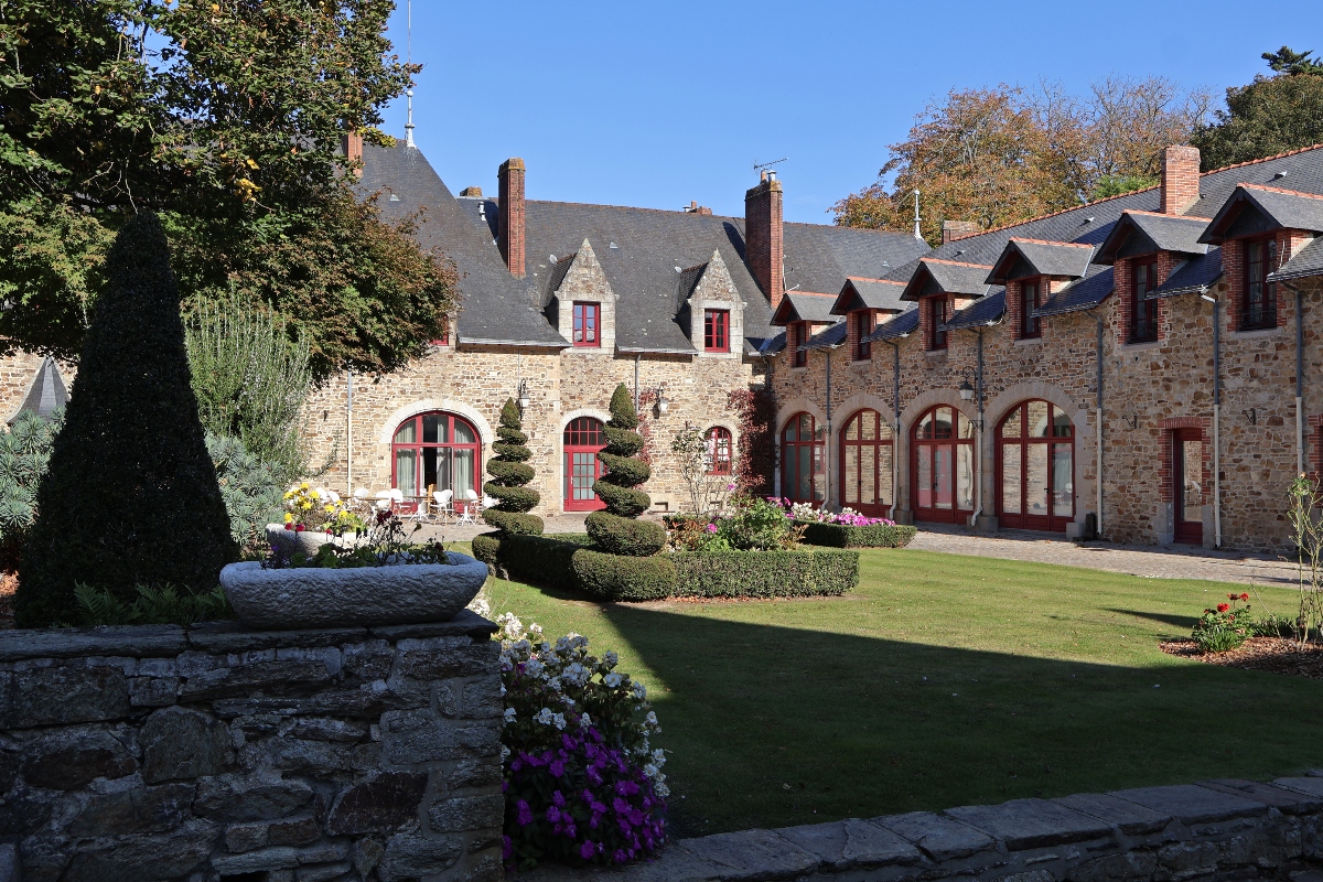 Stone courtyard buildings with grey slate roofs and distinctive red-trimmed arched windows and doors, featuring traditional French architecture with exposed stonework and dormer windows. The manicured grounds include a green lawn, sculpted topiary trees, colorful flower beds with purple and white blooms, and mature deciduous trees displaying autumn foliage under a clear blue sky.