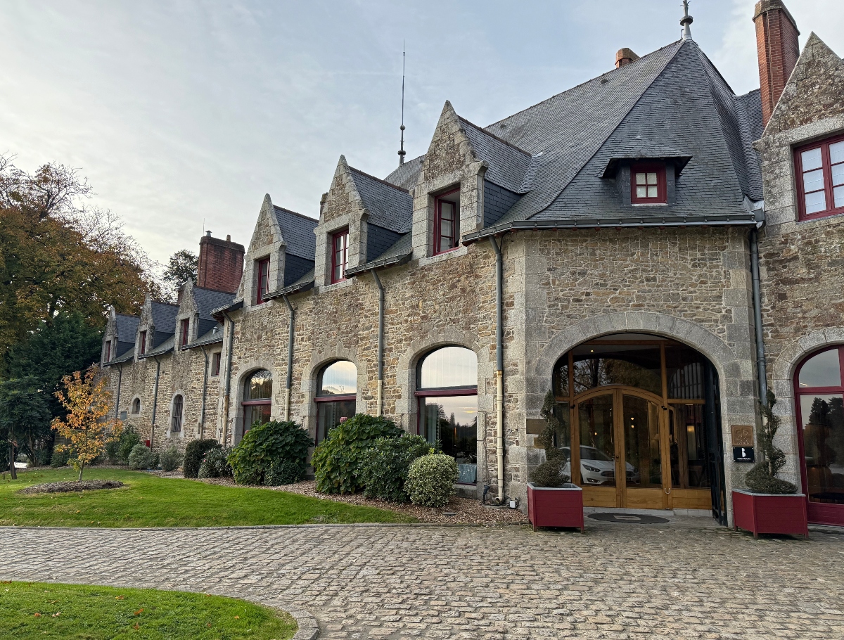 Stone manor building with weathered beige walls, dark slate roof with multiple pointed dormer windows, and burgundy-trimmed arched entrances on the ground floor. The entrance features wooden doors flanked by manicured topiary bushes, with a cobblestone courtyard and green lawn leading to the building under an overcast sky.
