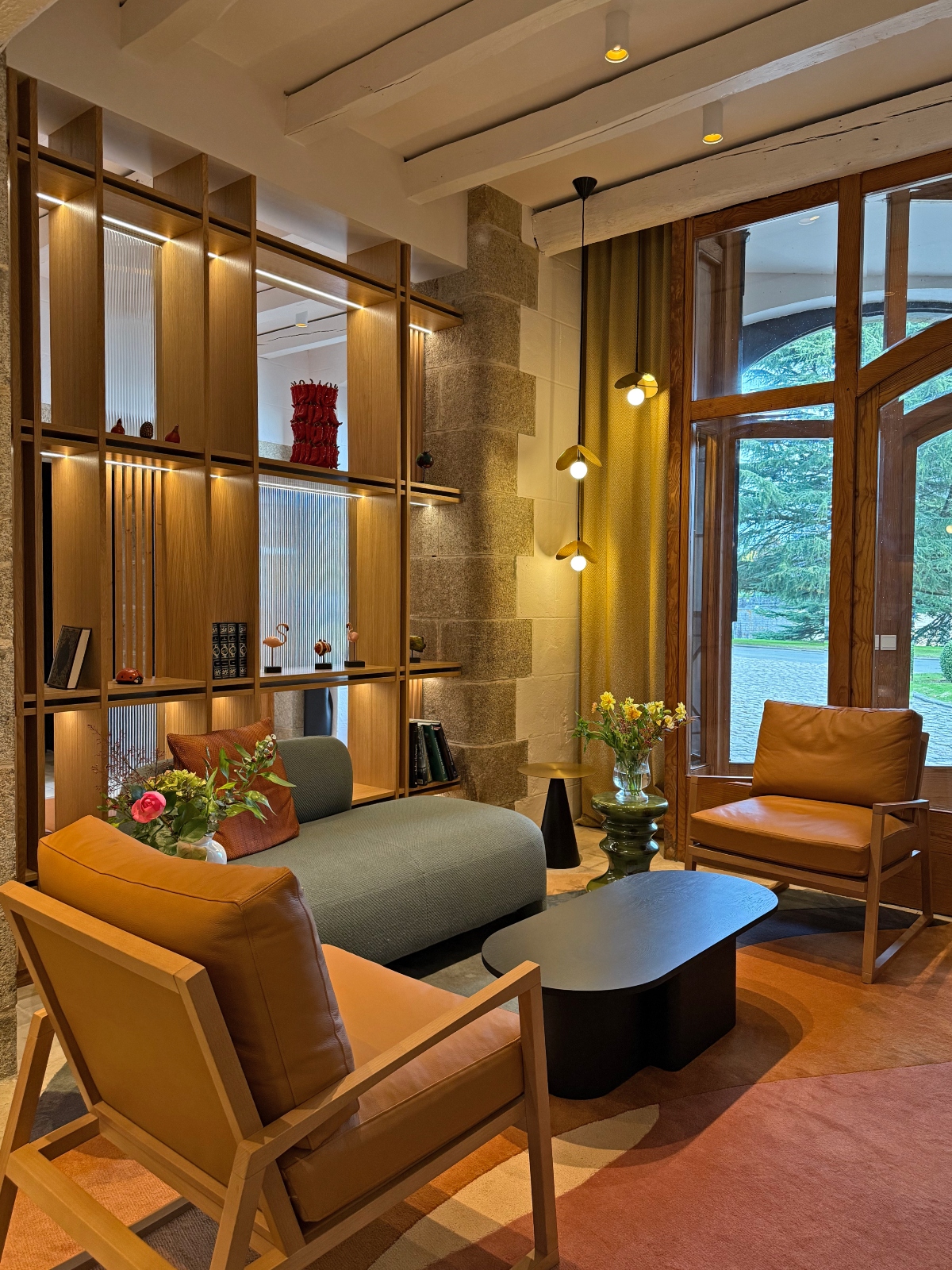 Modern hotel lobby featuring exposed stone walls, mid-century style furniture including tan leather armchairs and a gray sofa arranged around a black sculptural coffee table, with globe pendant lights suspended from the ceiling. The space includes custom wooden shelving with decorative objects, large wooden-framed windows overlooking grounds, and terracotta floor tiles accented with curved sections in rust, pink, and navy tones.