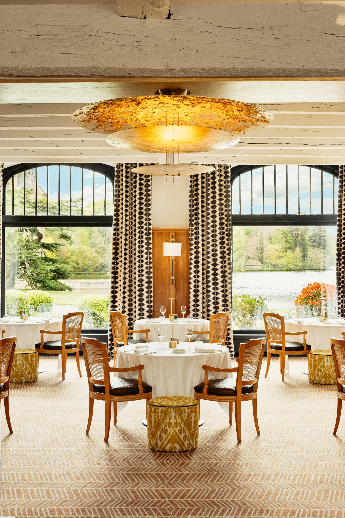 Fine dining restaurant interior with round tables dressed in white linens, wooden cane-back chairs with dark cushions, and a large circular amber-toned pendant light fixture suspended from an exposed beam ceiling. The space features tall arched windows with black frames overlooking a lake and landscaped grounds, geometric-patterned curtains, gold ikat-upholstered ottomans, and beige herringbone-patterned carpet flooring.