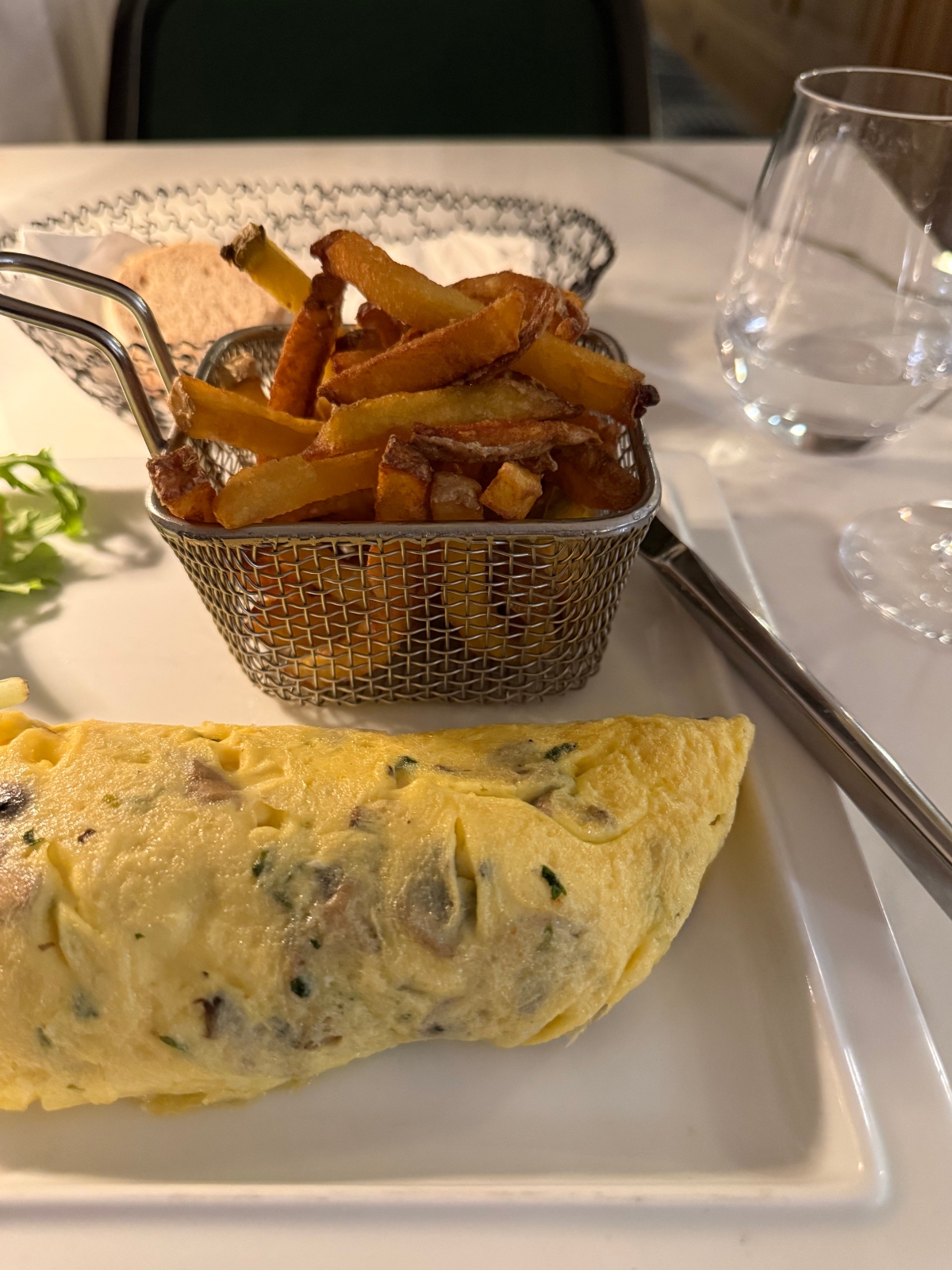 White rectangular plate with a folded herb-flecked omelet and a wire mesh basket containing golden-brown french fries, accompanied by a water glass. Additional plates with bread and garnishes visible in the background on a white tablecloth.