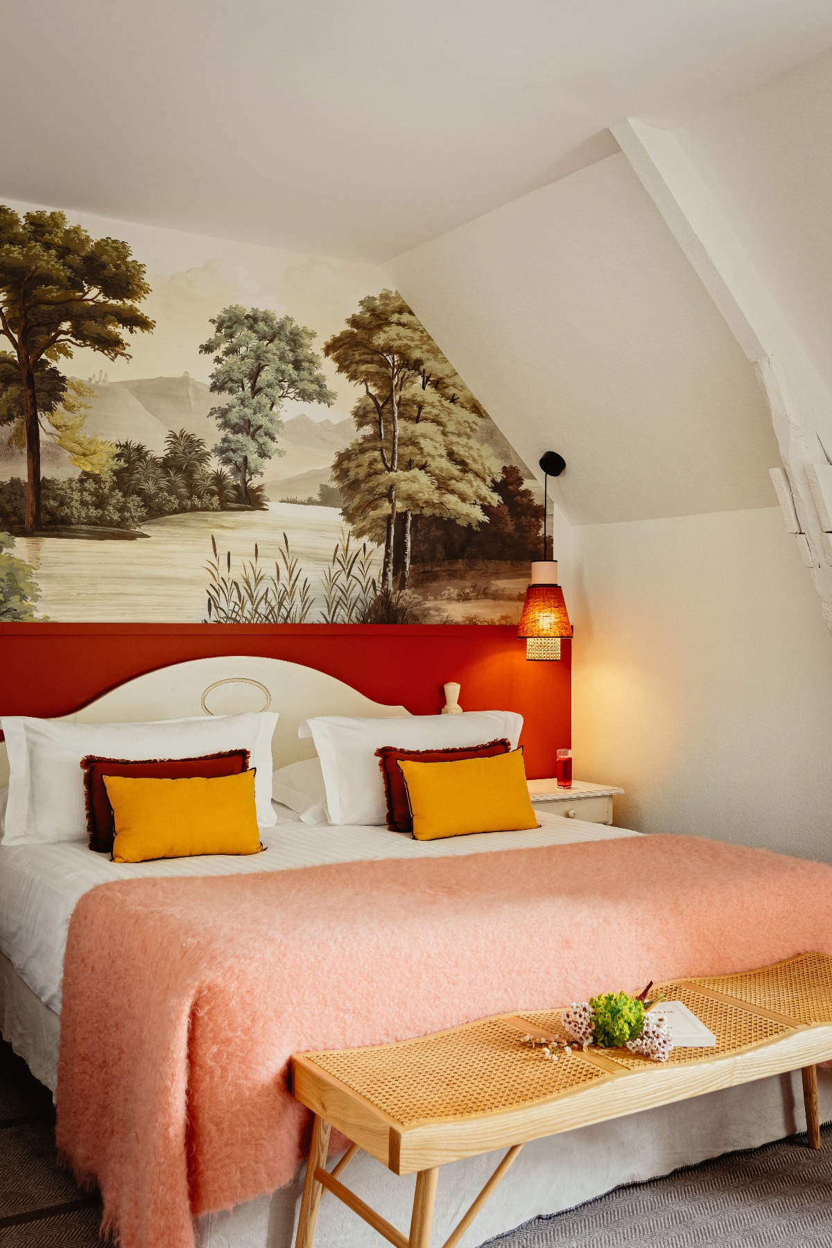 Bedroom with two white carved headboards against a rust-red accent wall topped by a landscape mural depicting trees and water in sepia tones, featuring white bedding with golden yellow and burgundy pillows and a pink throw blanket. The space includes sloped white ceilings, a woven cane bench at the foot of the bed with fresh flowers, and an orange pendant light fixture.