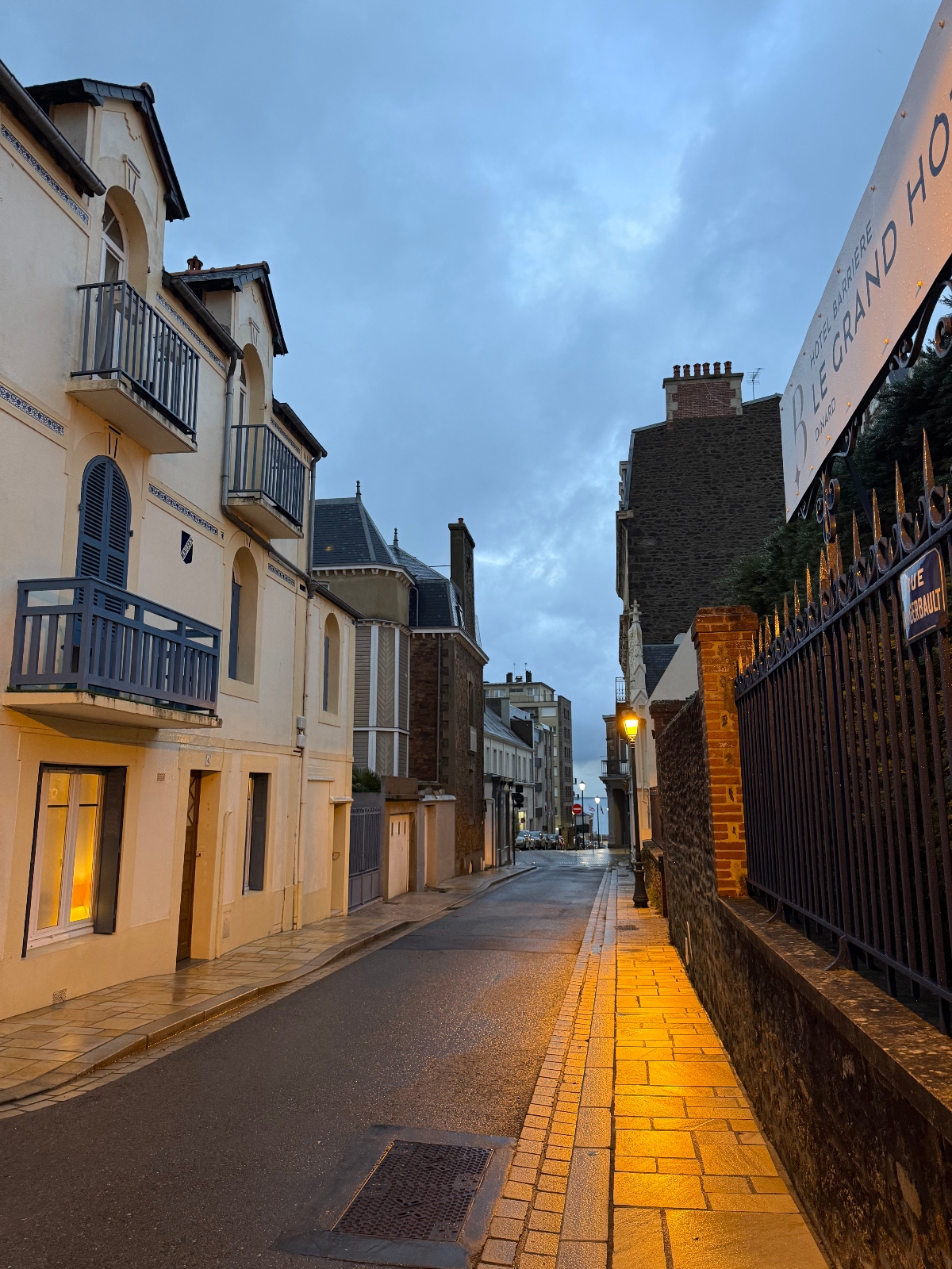 A narrow street at dusk lined with mixed-era buildings including a pale yellow villa with blue balconies on the left and darker stone buildings on the right, with a partially cloudy sky above. Streetlights illuminate an orange tile sidewalk beside the empty roadway, which curves gently into the distance where more buildings are visible.