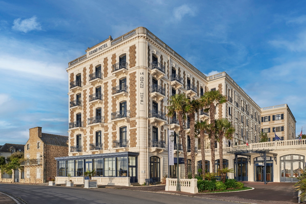 The exterior facade of Le Grand Hotel, a multi-story Belle Époque building with distinctive horizontal bands of cream stone and red brick, featuring wrought-iron balconies on each floor and the hotel name displayed vertically on the corner tower. Palm trees stand in front of the hotel's modern glass-enclosed ground floor entrance, with white stone balustrades framing the forecourt under a clear blue sky.
