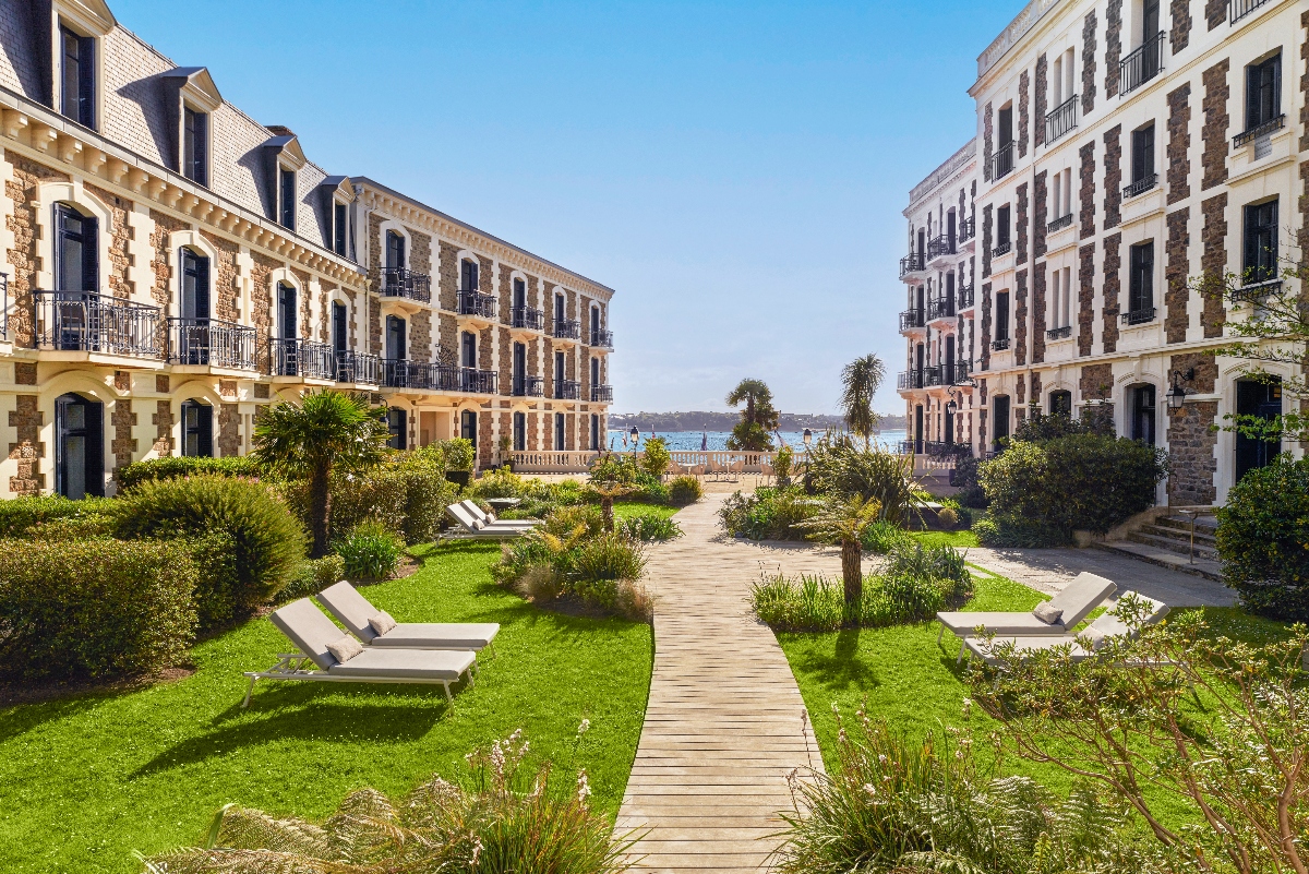 A wooden boardwalk pathway runs through a landscaped garden with manicured lawns, lounge chairs, and Mediterranean plantings including palm trees and shrubs, flanked by matching Belle Époque hotel buildings with cream stone and red brick facades. The garden opens to a view of the harbor beyond a white stone balustrade under a clear blue sky.