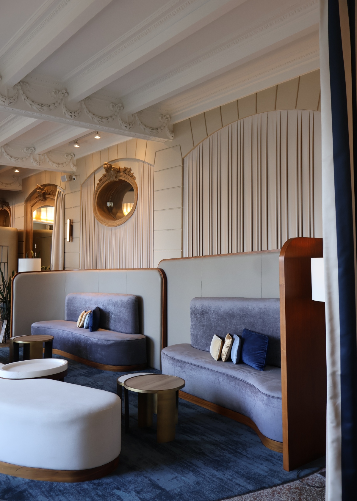 A hotel lobby with high ceilings featuring ornate white molding, cream-colored vertical wall paneling, and a large circular mirror with an elaborate gold frame mounted on the wall. The seating area includes curved blue velvet banquettes with navy accent pillows, paired with white round ottomans and dark cylindrical side tables with brass accents on a navy area rug.