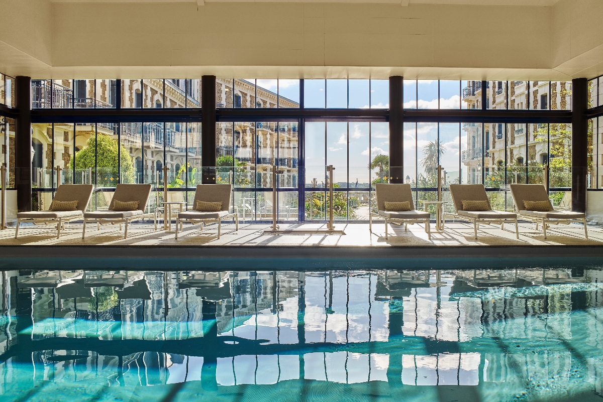 An indoor swimming pool with turquoise water in the foreground, with a row of tan lounge chairs arranged along the deck beneath a beige ceiling. Floor-to-ceiling windows and black structural columns frame views of the hotel's Belle Époque facade with its balconies, along with palm trees and landscaping visible outside.