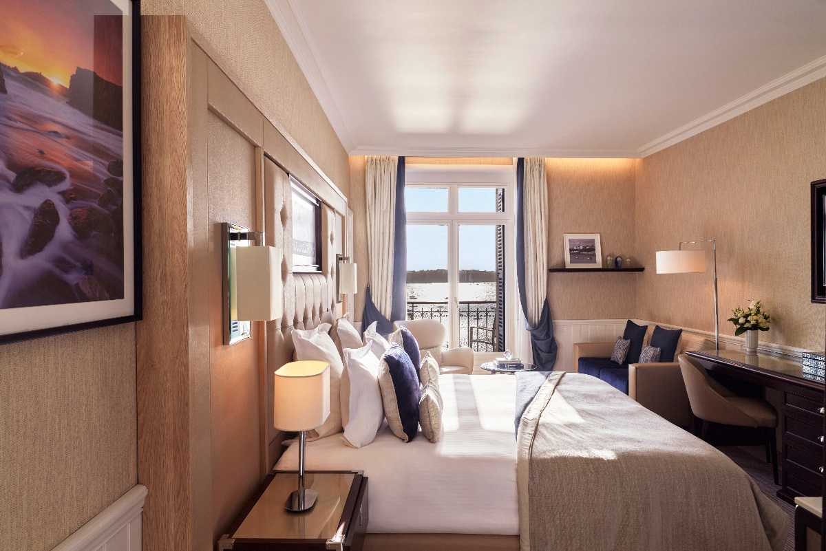 A hotel bedroom featuring a bed with a tufted upholstered headboard and white linens accented with navy pillows, positioned against a wall with light wood and beige paneling. French doors with cream curtains open to a balcony with an iron railing overlooking water, with a writing desk and floor lamp positioned to the right of the windows.