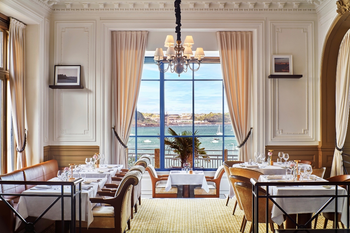 A formal restaurant dining room with white paneled walls, ornate crown molding, and peach-colored curtains framing a large central window overlooking a harbor with boats and a distant shoreline. Multiple tables are set with white linens and wine glasses, arranged beneath a traditional multi-arm chandelier, with upholstered dining chairs in caramel and cream tones positioned around each table.