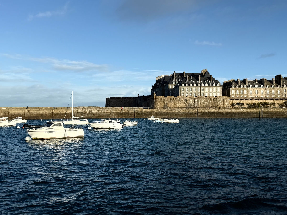 Several white motorboats moored in dark blue harbor water in the foreground, with a massive stone fortification wall rising behind them. Historic multi-story buildings with gray slate roofs and dormer windows line the top of the ramparts under a blue sky with scattered clouds.