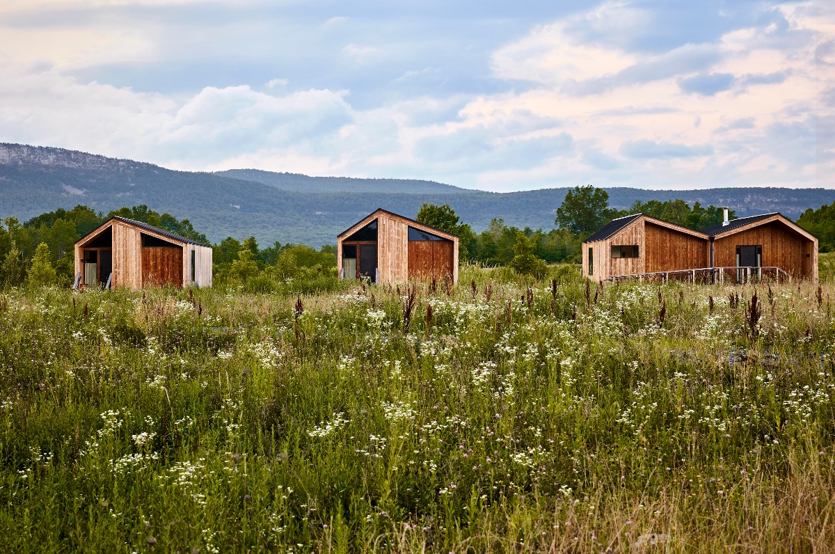 Three modern wooden cabins with peaked roofs and large windows are positioned in a row across a wildflower meadow filled with white blooms and tall grasses. Mountain ridges covered with trees form the backdrop beneath a blue sky with white clouds.