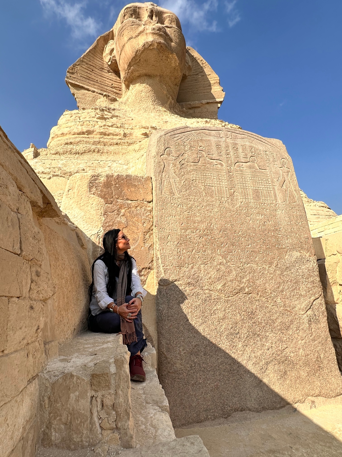 Woman in casual clothing sits on stone steps looking up at the Great Sphinx of Giza, photographed from between its massive front paws. A large limestone stela covered with hieroglyphic inscriptions stands vertically against the sphinx's chest, flanked by weathered stone walls under a blue sky.
