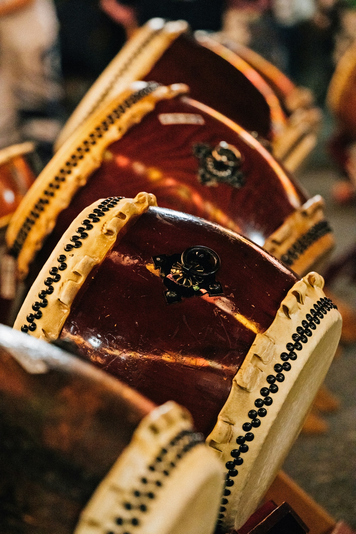 Traditional Japanese taiko drums with dark reddish-brown lacquered bodies and light-colored rope rims secured with rows of black metal tacks. Japanese characters are visible on the drum faces, and multiple drums are arranged in the frame with shallow depth of field creating a blurred background.