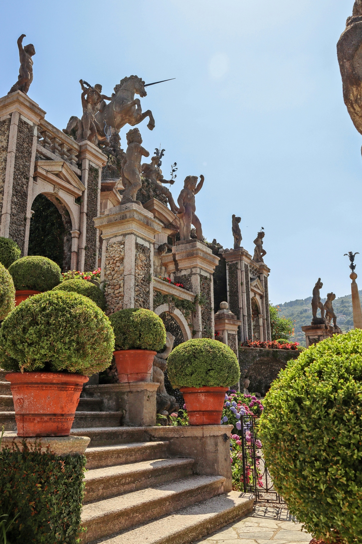 A baroque terraced garden features stone steps lined with large terracotta pots containing rounded topiary shrubs and colorful flowering plants. Multiple classical statues, including a prominent equestrian figure at the top, are positioned on tiered stone platforms decorated with pebble mosaics and arched architectural elements, with a hillside visible in the background.
